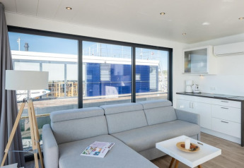 Sala de estar moderna en una casa flotante, sofá gris claro, pequeña cocina y grandes ventanales al muelle.