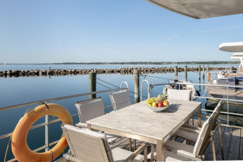 Udsigt fra husbåd med terrassebord, stole og frugtskål, beliggende ved rolig havn under klar himmel.