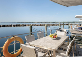 Terras van woonboot met tafel, stoelen en fruitschaal, uitkijkend op rustige haven bij helder weer.