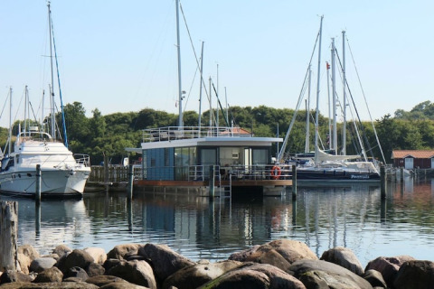 A modern houseboat docked in a marina among sailboats with trees and rocky shore in the background.