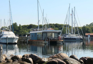 Een moderne woonboot aangemeerd in een haven tussen zeiljachten met bomen en rotsen rondom.