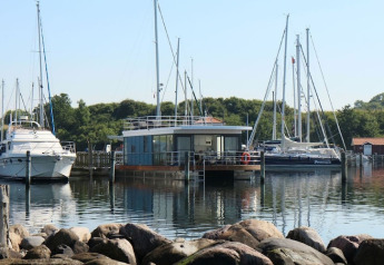 Een moderne woonboot aangemeerd in een haven tussen zeiljachten met bomen en rotsen rondom.
