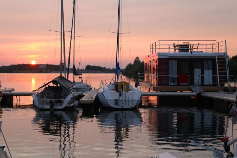 Solnedgang over en husbåd og sejlbåde ved en rolig havn, spejlet i vandet med bløde farver i himlen.