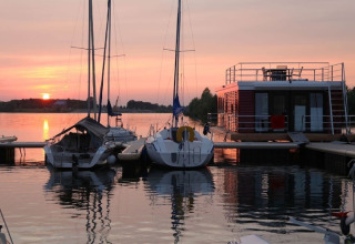 Houseboat and sailboats docked at a tranquil marina during sunset, calm water reflecting the colorful sky.