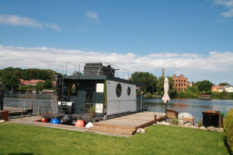 Il moderno houseboat Friedrich è ormeggiato su un fiume, con prato e area relax in primo piano.
