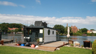 The modern Houseboat Friedrich is moored by a river, with a grassy yard and seating area in the foreground.