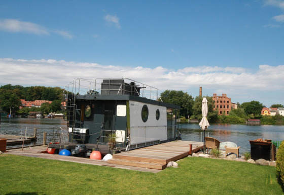 Le Houseboat Friedrich moderne est amarré au bord d'une rivière, avec pelouse et coin détente au premier plan.