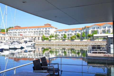 Udsigt fra en husbåd med stole, marina med lystbåde og moderne bygninger i baggrunden under blå himmel.