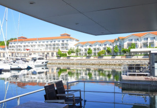 Vista da una casa galleggiante con sedie, marina, yacht e moderni edifici sul lungomare sotto un cielo sereno.