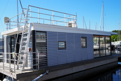 Moderne husbåd med tagterrasse og metalrækværk, fortøjet i en marina med sejlbåde i baggrunden.
