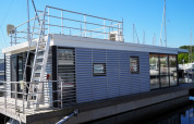 Modern houseboat with rooftop deck and ladder, docked at a marina with sailboats in the background.