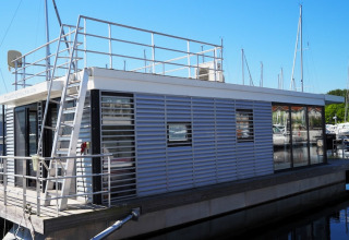 Moderne husbåd med tagterrasse og metalrækværk, fortøjet i en marina med sejlbåde i baggrunden.