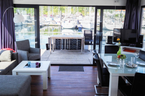 Moderne stue på en husbåd med udsigt over en marina, store vinduer, spisebord og udendørs terrasse.