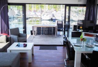 Moderne stue på en husbåd med udsigt over en marina, store vinduer, spisebord og udendørs terrasse.