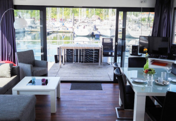 Modernes Wohnzimmer auf einem Hausboot mit Blick auf einen Jachthafen, großem Fenster und Terrasse.