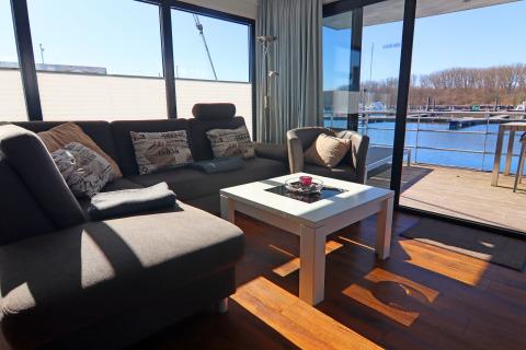 Soggiorno su una houseboat con divano grigio, ampie finestre e vista su terrazza e acqua all’esterno.