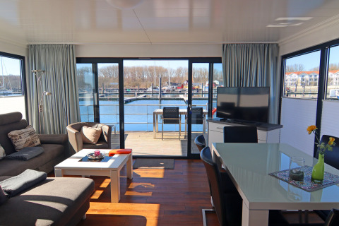 Moderne husbåd med lyst interiør, panorama vinduer samt terrasse med udsigt over en marina.