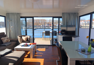 Moderne husbåd med lyst interiør, panorama vinduer samt terrasse med udsigt over en marina.