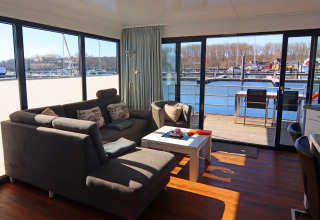 Stue med moderne sofa og stort vindue, der giver udsigt over vandet og en terrasse på en husbåd.