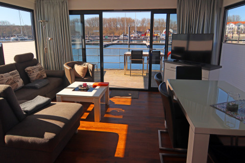 Gezellige woonboot met eigentijdse inrichting, eethoek, televisie en uitzicht op de jachthaven via grote ramen.