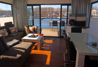Gezellige woonboot met eigentijdse inrichting, eethoek, televisie en uitzicht op de jachthaven via grote ramen.