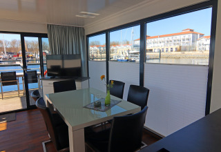 Moderne stue på husbåd med store vinduer, udsigt til marina i Tyskland, spisebord og TV-område.