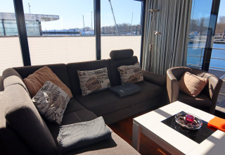 Hyggelig stue på husbåd Family & Friends LP2 i Tyskland med store vinduer, sofa og udsigt til havnen.