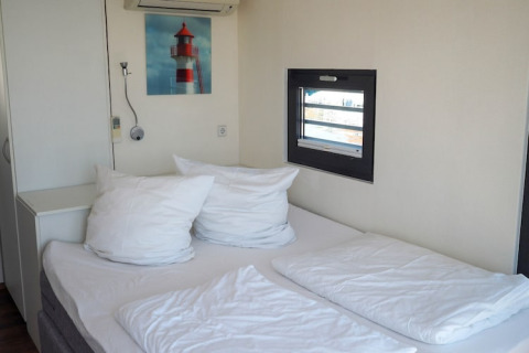 Slaapkamer op een woonboot met dubbel bed, wit beddengoed, klein raam en een schilderij van een vuurtoren.
