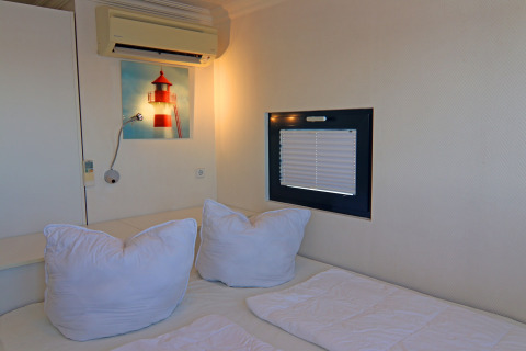 Camera da letto su una houseboat con biancheria bianca, piccola finestra e quadro di un faro al muro.