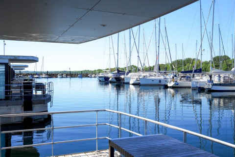 Zicht vanaf een woonboot met houten tafel en leuning op een jachthaven vol zeilboten bij zonnig weer.