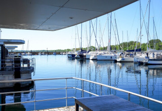 Zicht vanaf een woonboot met houten tafel en leuning op een jachthaven vol zeilboten bij zonnig weer.