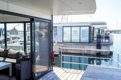Moderne husbåde med store vinduer og terrasse på vandet, set fra dækket på en af husbådene i en havn.