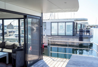 Moderne husbåde med store vinduer og terrasse på vandet, set fra dækket på en af husbådene i en havn.