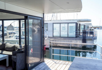 Casa flotante moderna con ventanales y terraza sobre el agua, vista desde otra casa flotante en el puerto.