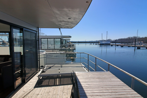 Udsigt fra en husbåd terrasse ud over marinaen med klart vand, både og blå himmel i baggrunden.