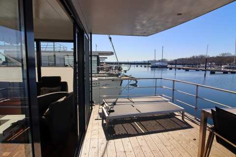 Uitzicht vanop het terras van een woonboot met ligbed over het water, boten en een zonnige jachthaven.
