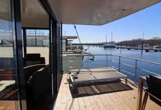 Uitzicht vanop het terras van een woonboot met ligbed over het water, boten en een zonnige jachthaven.