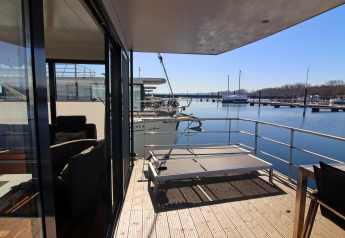 View from a houseboat patio with sunbed overlooking water, boats, and a marina on a bright sunny day.