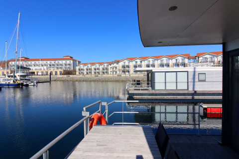 Udsigt fra en husbåd med terrasse over marinaen, moderne bygninger og både i baggrunden under blå himmel.