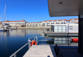 Uitzicht vanaf het dek van een woonboot in een haven met moderne gebouwen en boten bij heldere lucht.