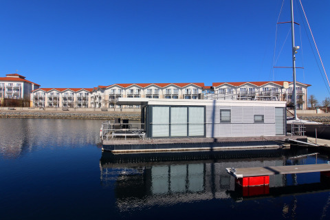 Husbåd fortøjet i en rolig havn med moderne lejlighedsbygninger i baggrunden under en klar blå himmel.