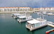 Moderne woonboten aangemeerd in een haven met appartementencomplexen op de achtergrond en bewolkte lucht.