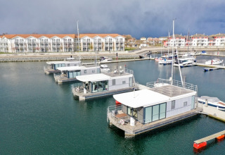 Houseboat moderne ormeggiate in un porto, con appartamenti di stile sullo sfondo e cielo nuvoloso.