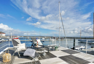 Afslappende terrasse på en husbåd med liggestole, bord og udsigt over vandet og bådehavnen.