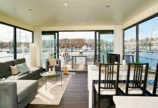 Salon lumineux sur une péniche avec grandes fenêtres, table à manger et vue sur le port de plaisance.
