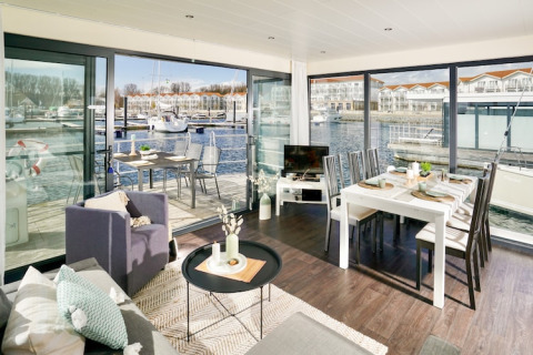 Sala moderna en una casa flotante con comedor, grandes ventanales y vista a la marina con yates al sol.