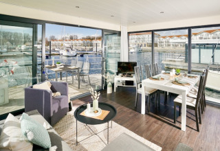 Zona giorno moderna su houseboat con tavolo da pranzo, ampie vetrate e vista marina con yacht al sole.