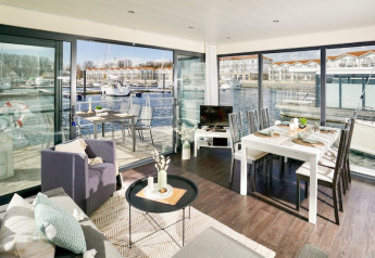 Modern houseboat living area with dining table, big windows and marina view with yachts in sunny weather.