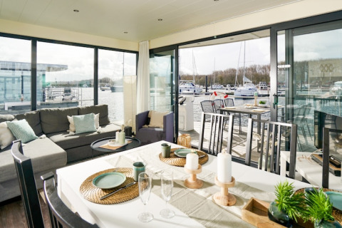 Moderne leefruimte en eetkamer op een woonboot met zicht op jachthaven en zeilboten buiten.