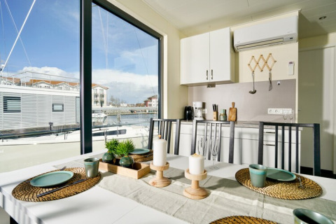 Table à manger sur une péniche avec vue sur le port et les bateaux, grandes fenêtres et cuisine moderne.